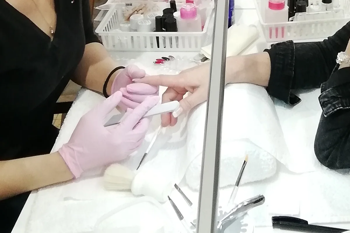 Vorbereitung des Naturnagels im Nageldesign-Kurs: Eine Teilnehmerin mattiert mit Handschuhen und Feile den Nagel ihrer Partnerin, daneben Pinsel, Instrumente und Materialkorb.