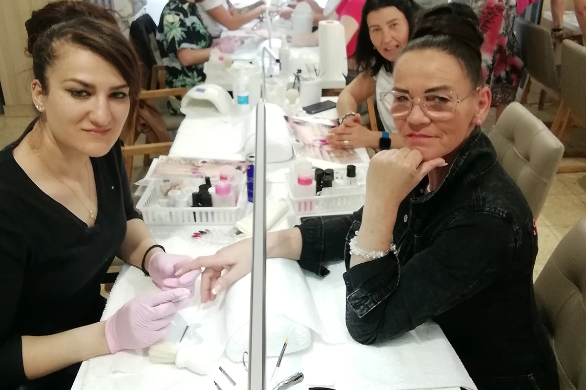 Teilnehmerinnen einer Nageldesign-Ausbildung arbeiten paarweise am Schulungstisch: eine Teilnehmerin modelliert mit Handschuhen an der Hand ihrer Partnerin, daneben LED-Lampe, Feilen und Materialkörbe.