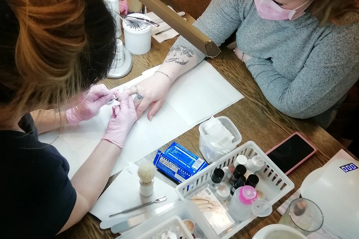 Nahaufnahme einer Nagelmodellage im Kurs: Eine Teilnehmerin arbeitet mit Handschuhen und Pinsel am Naturnagel ihrer Partnerin, daneben Feilen, Gele und Desinfektionsmittel.
